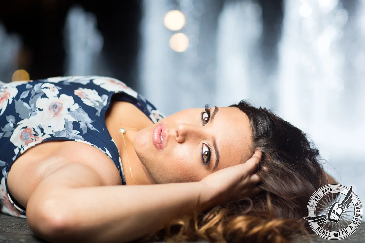 Woman in a floral dress lying on her side with one hand in her dark hair, looking toward the camera.