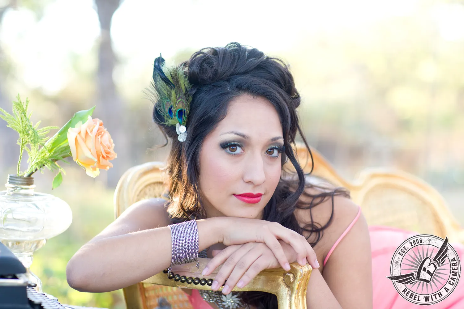 Woman in a pink dress with a peacock-feather hair accessory leaning on an ornate gold chair beside a vase with a peach rose.