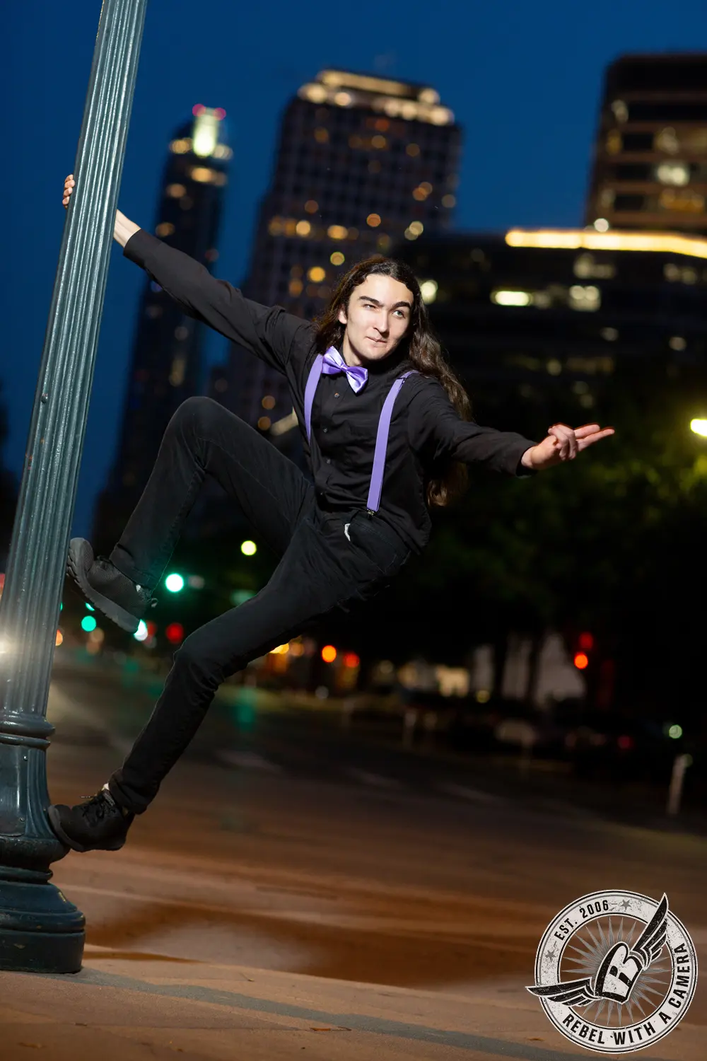 Alexander in black clothing with purple suspenders and bow tie, holding onto a lamppost at night with city buildings behind him.