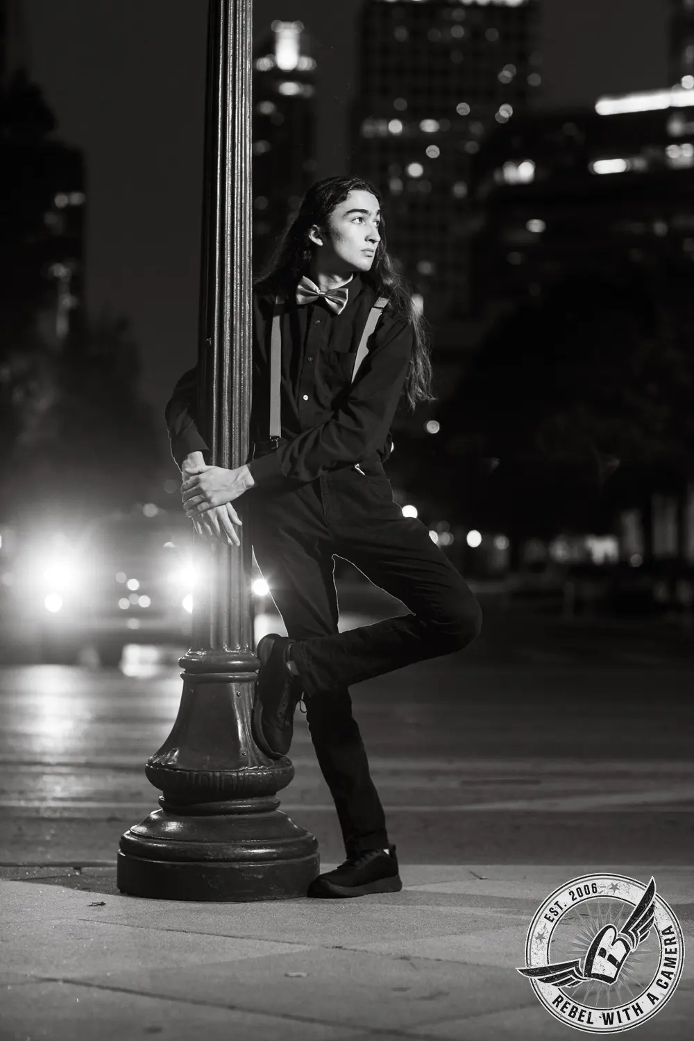 Black-and-white nighttime portrait of Alexander leaning against a decorative streetlamp, wearing a bow tie and suspenders.