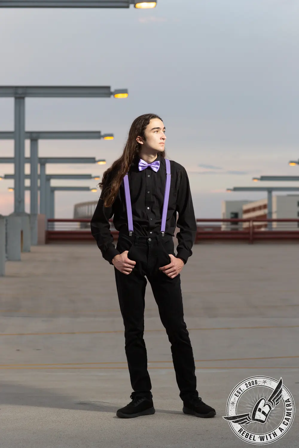 Alexander standing in a parking garage wearing a black outfit with purple suspenders and a purple bow tie.