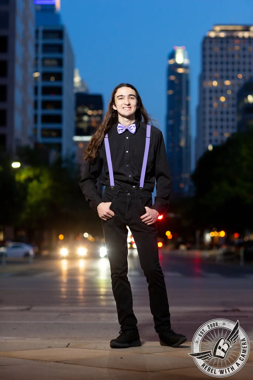 Alexander stands on a downtown sidewalk in a black shirt with purple suspenders and bow tie, with city buildings and traffic lights blurred behind him.