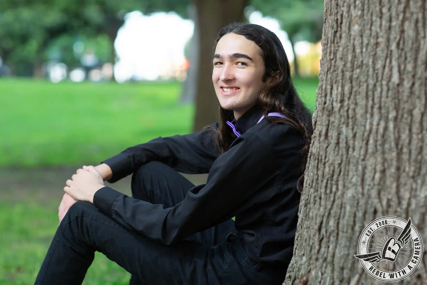 Alexander, wearing black, sits beside a tree in a green park and smiles toward the camera.