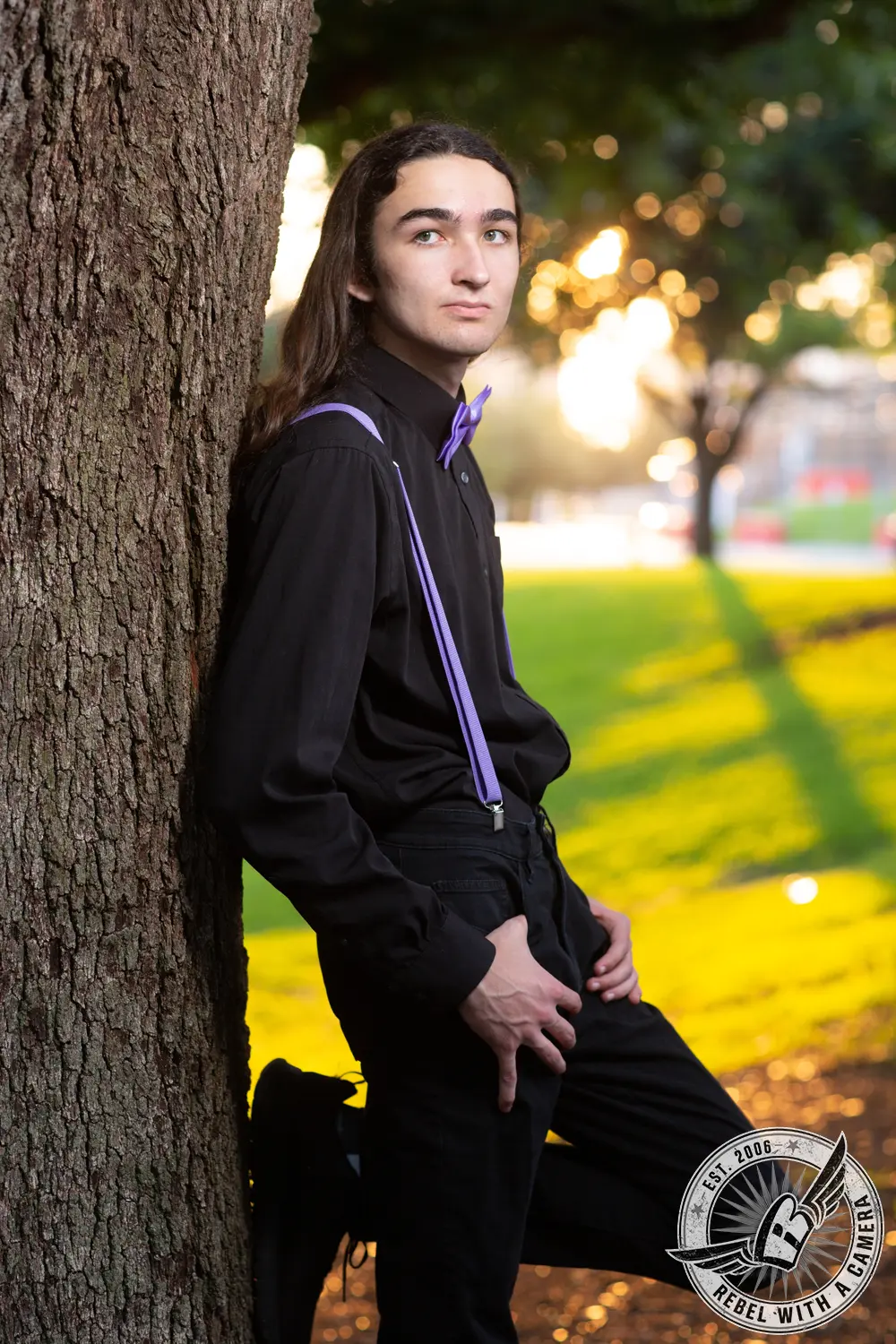Alexander leans against a large tree in a black shirt with purple suspenders and bow tie, with a sunlit park behind him.