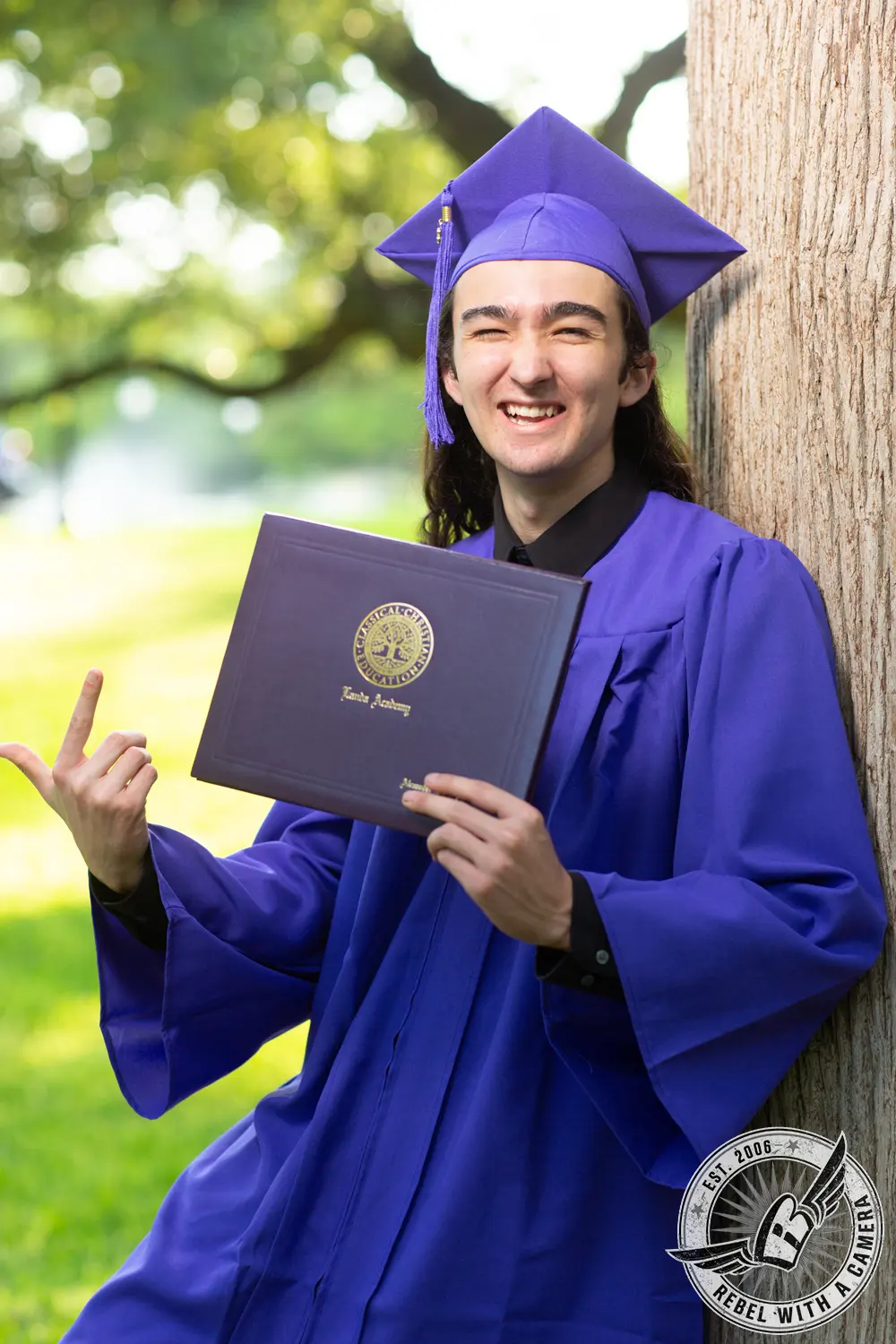 Alexander in a purple graduation cap and gown holds a diploma while leaning against a tree.