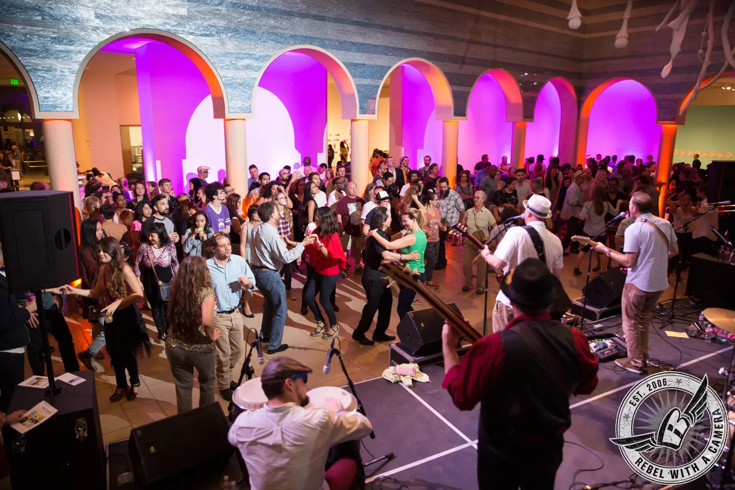 A large crowd gathers and dances in an arched museum space as a live band performs onstage under purple and orange lighting.