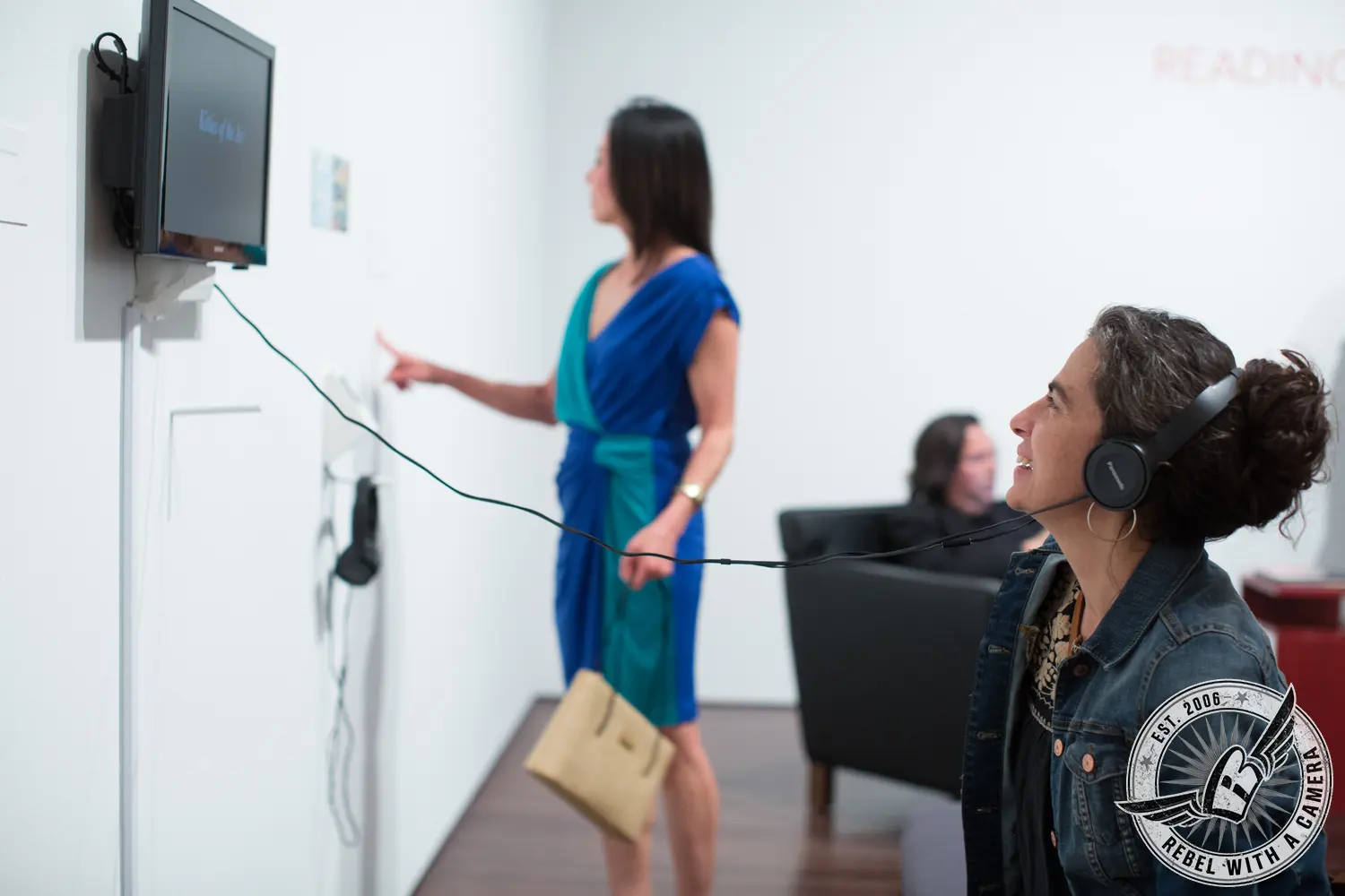 A guest wearing headphones looks toward a wall-mounted screen while another guest points at the display in a museum gallery.