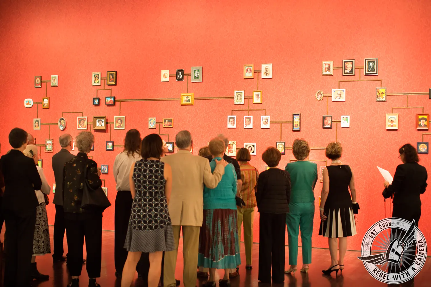 Director’s Circle guests stand before a red gallery wall filled with small framed portraits connected by thin lines.