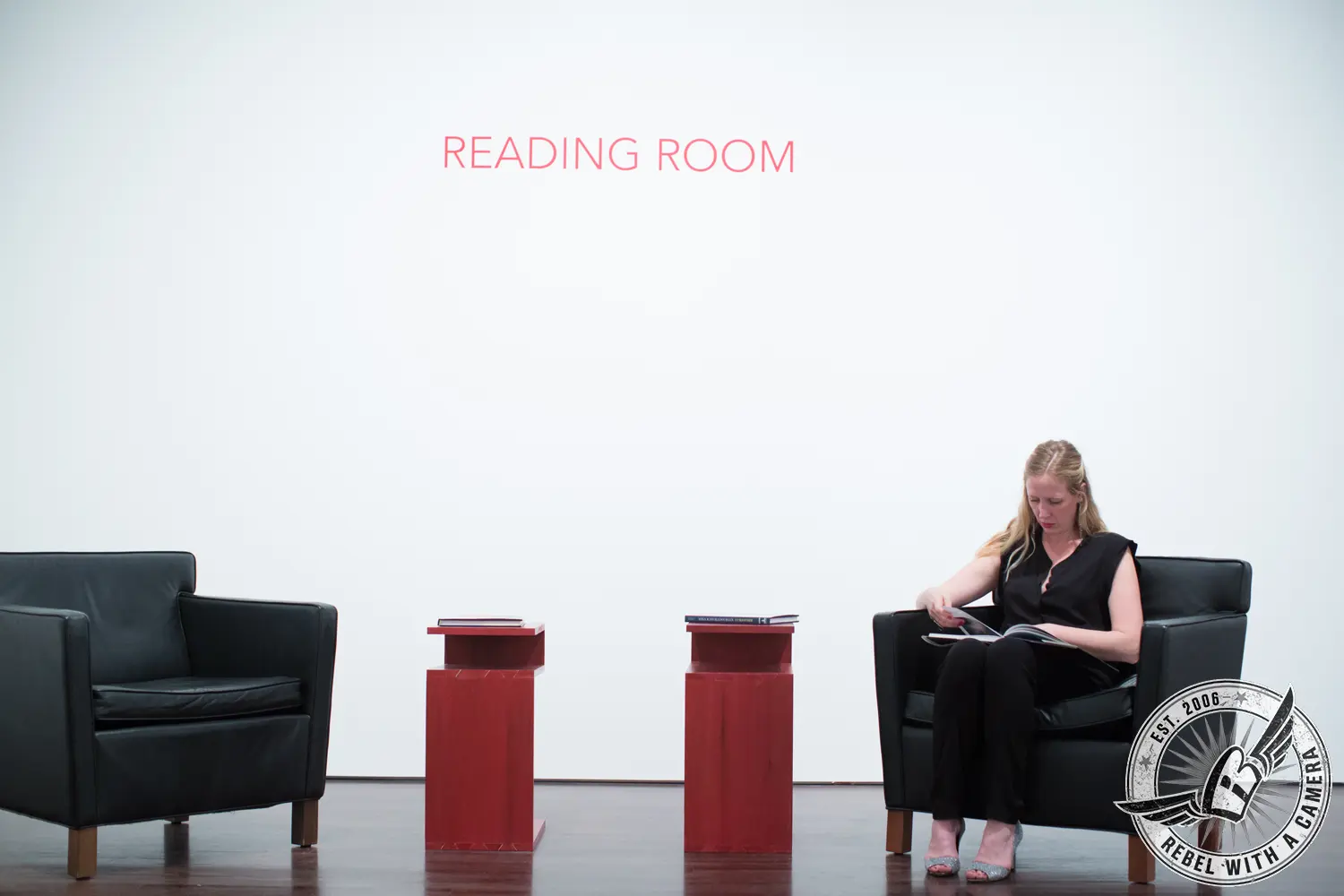 A woman in black sits in a black armchair reading a book beneath a wall sign that reads “READING ROOM.”