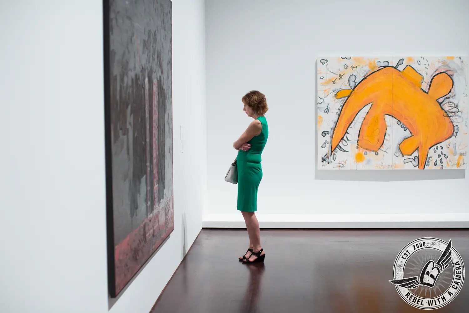 A woman in a green dress views a large dark painting in a white gallery, with a bright orange abstract artwork on the opposite wall.