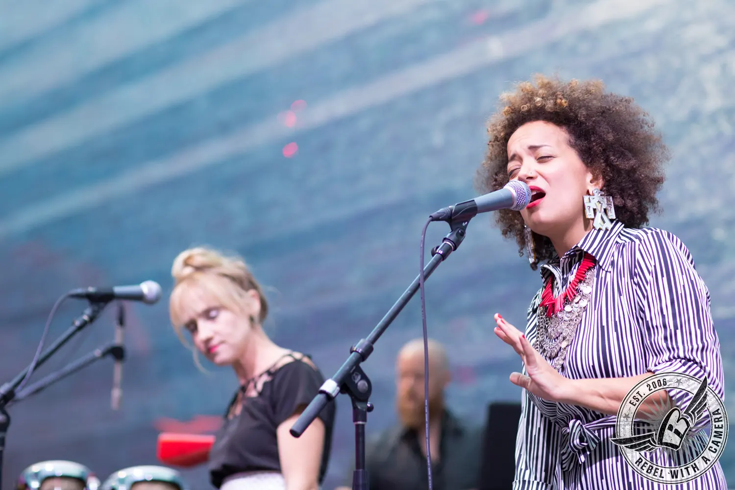 A singer in a black-and-white striped outfit performs into a microphone while another musician stands nearby.