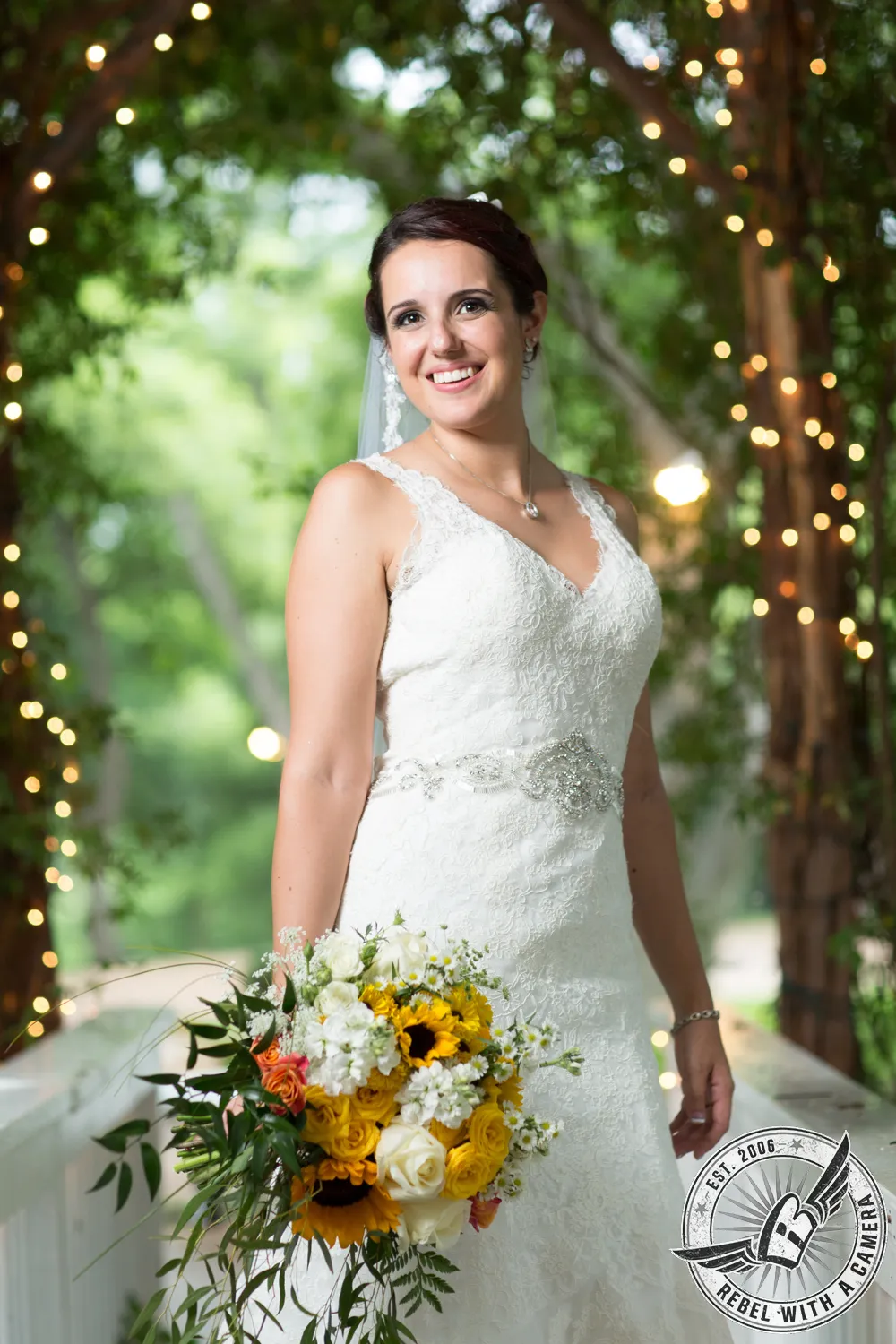 Bride holding a yellow and white bouquet beneath string lights