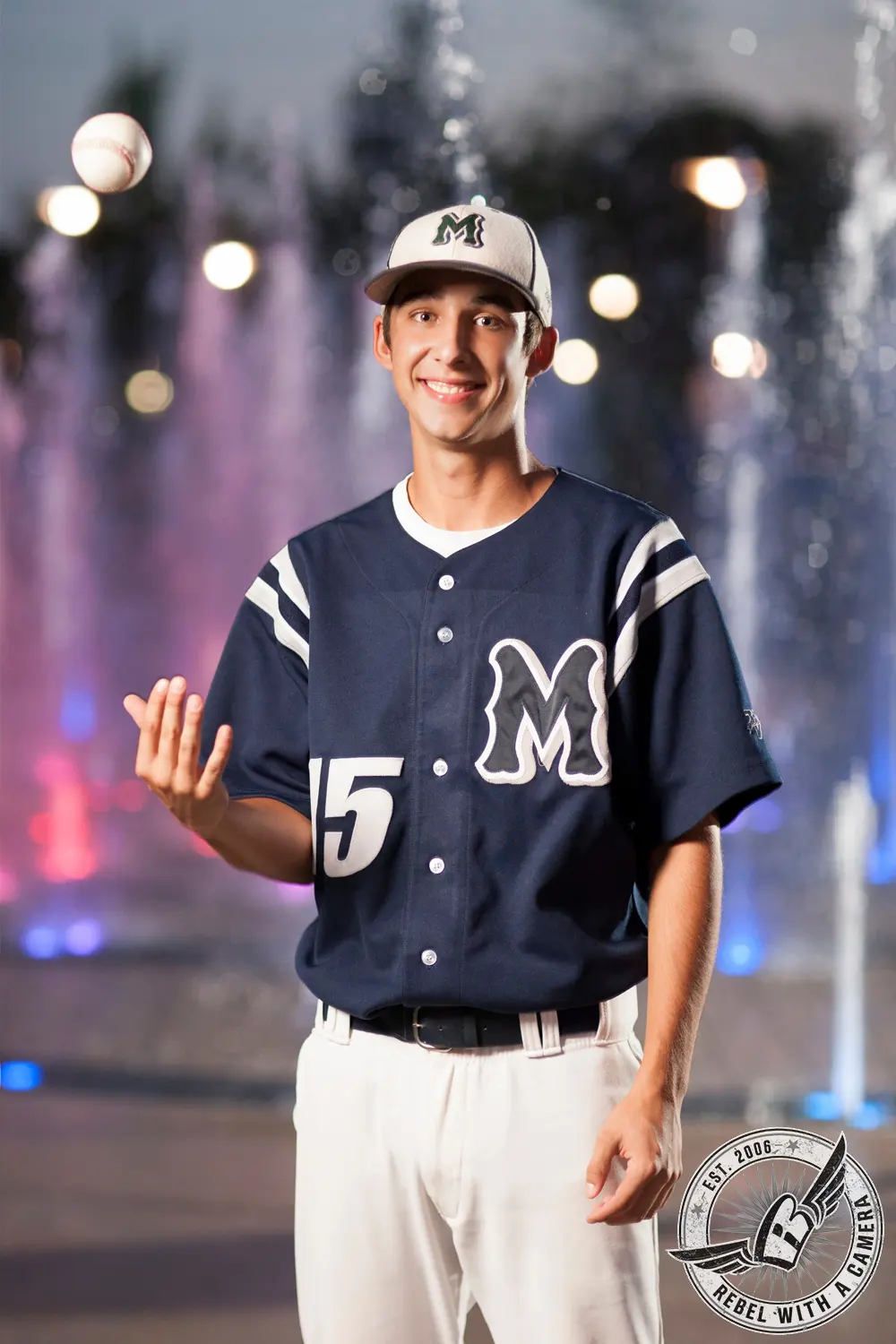 Jacob in a dark baseball uniform and cap tosses a baseball in front of illuminated fountains at night.