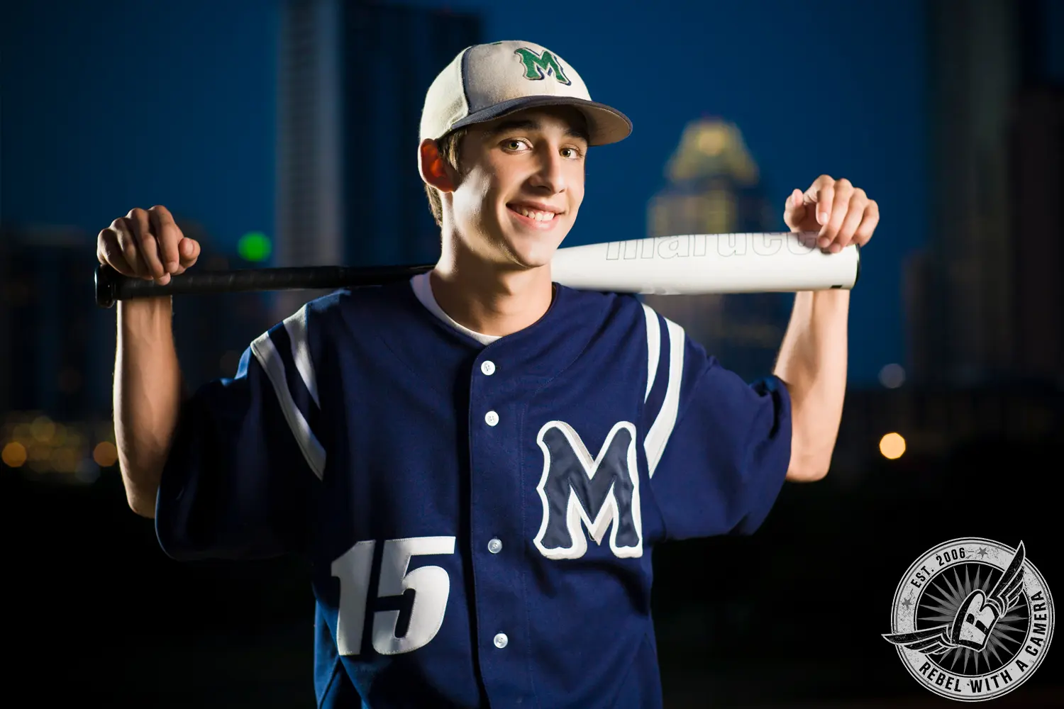 Jacob in a blue baseball uniform holds a bat across his shoulders during a nighttime senior portrait.