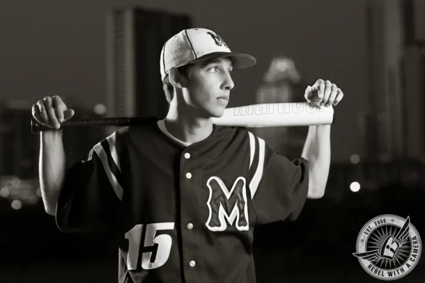 Black-and-white portrait of Jacob in a baseball uniform holding a bat across his shoulders at night.