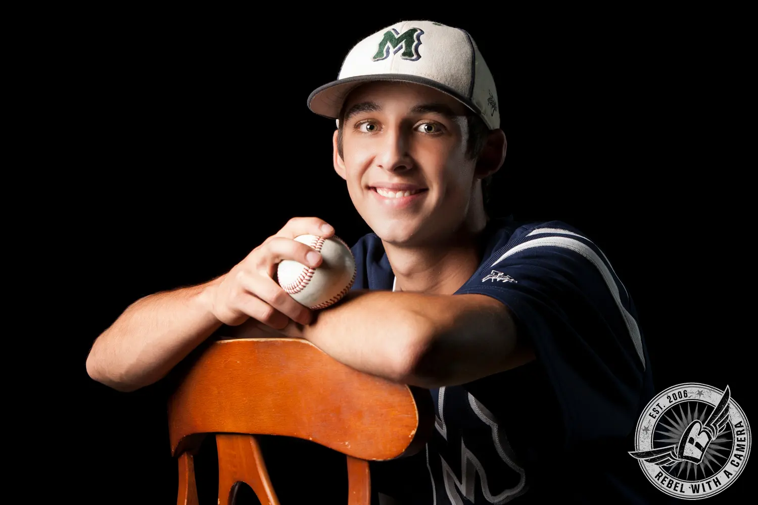 Jacob in a baseball uniform and cap holds a baseball while leaning on a wooden chair against a black background.