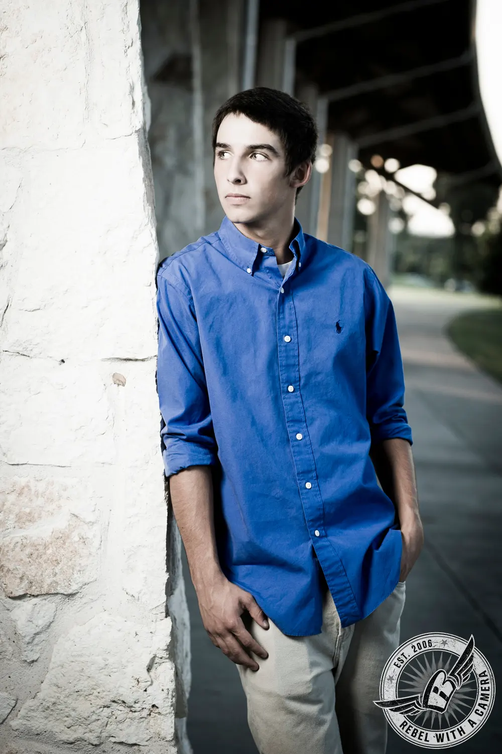 Jacob wearing a blue button-down shirt and light pants beside a textured white stone wall.