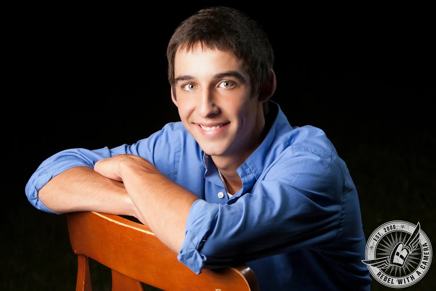 Jacob in a blue button-down shirt leaning on the back of a wooden chair against a black background.