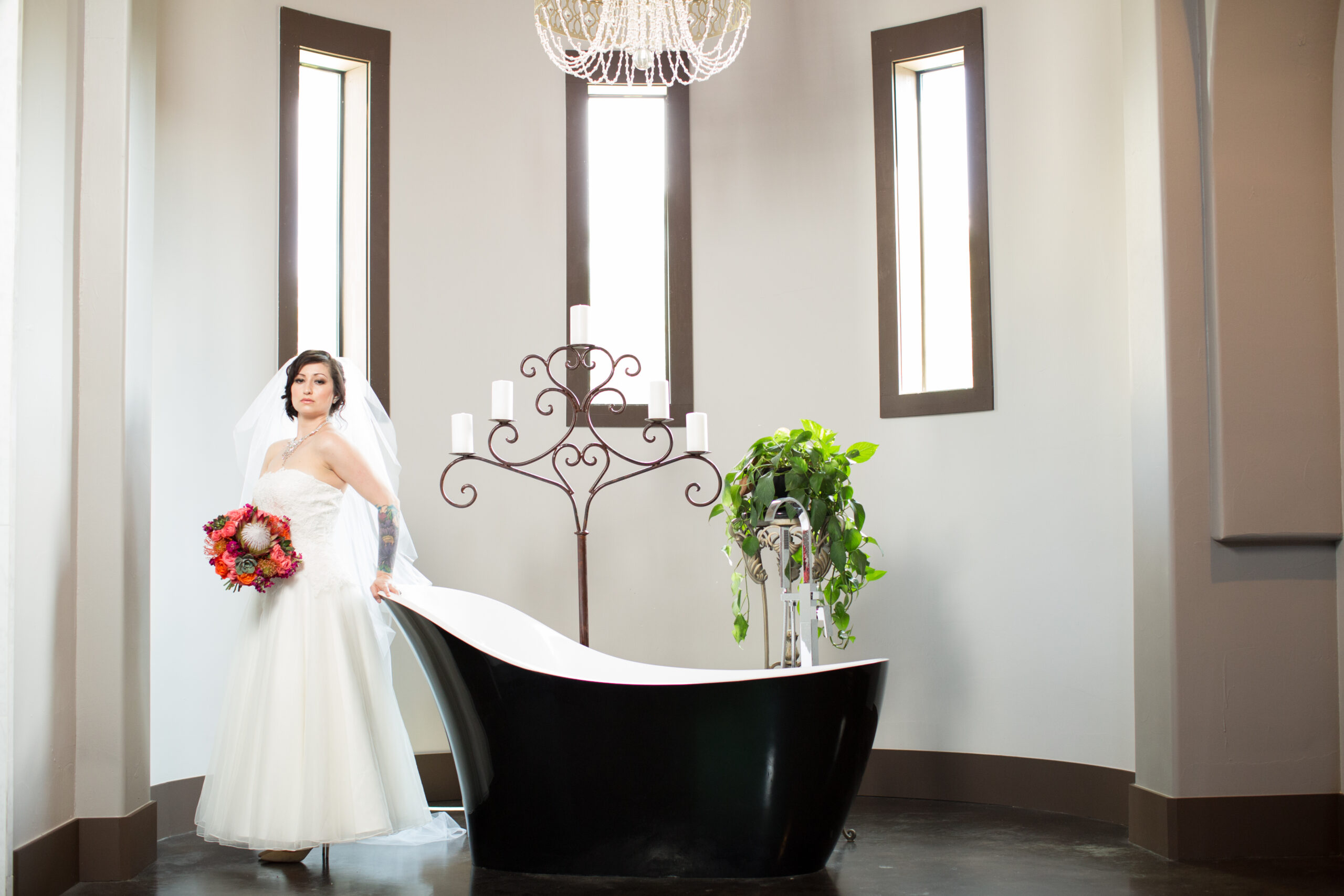 Bride in a white wedding dress and veil holding a colorful bouquet beside a black freestanding bathtub.
