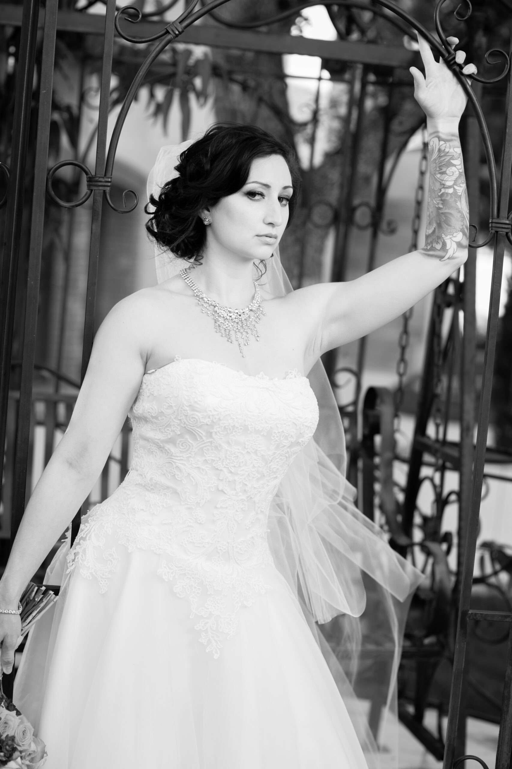 Black-and-white bridal portrait of Jessica in a lace wedding dress and veil, standing beneath an ornate wrought-iron arch.