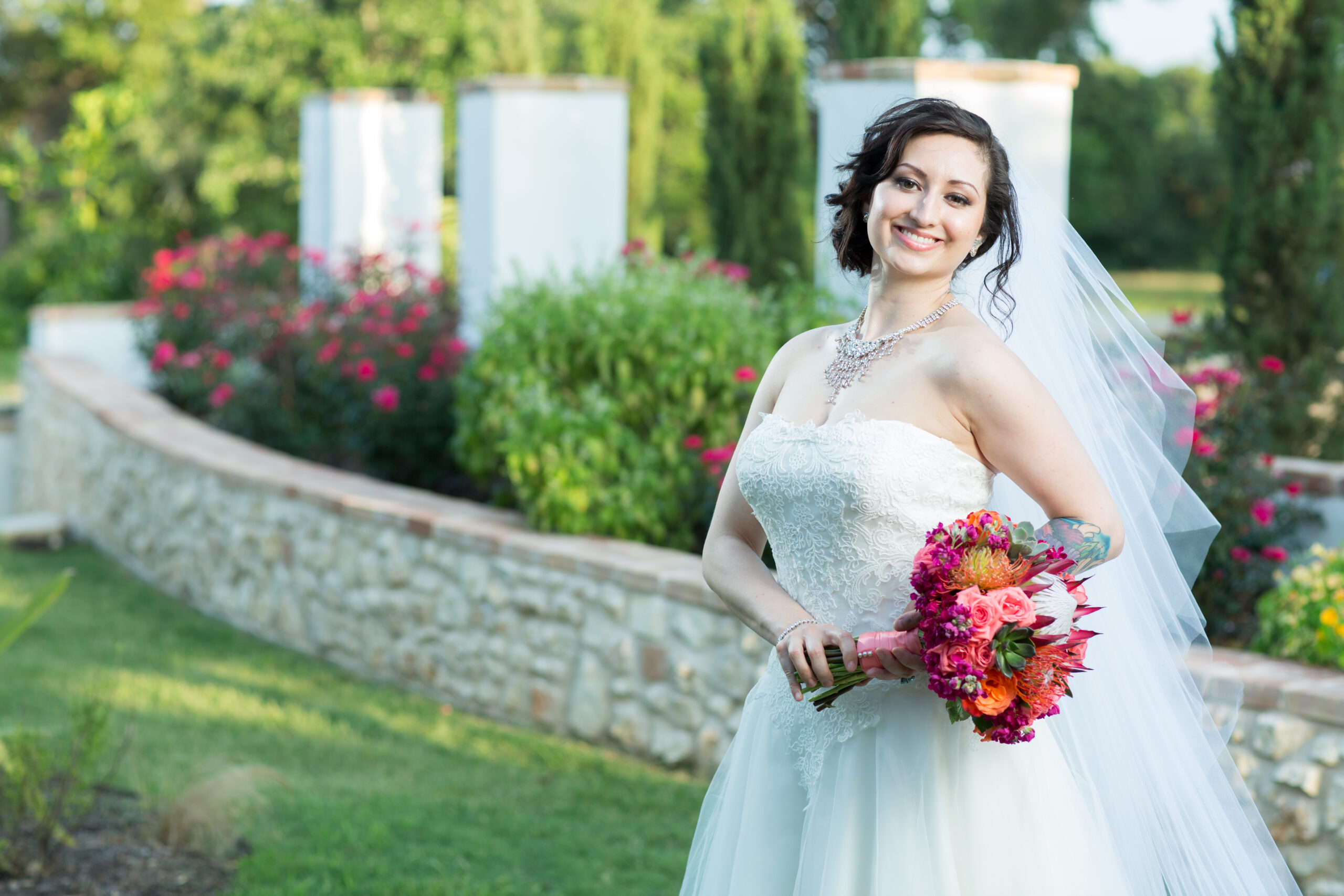 Bride in a white lace wedding dress and veil holding a vibrant pink and orange bouquet in a garden.