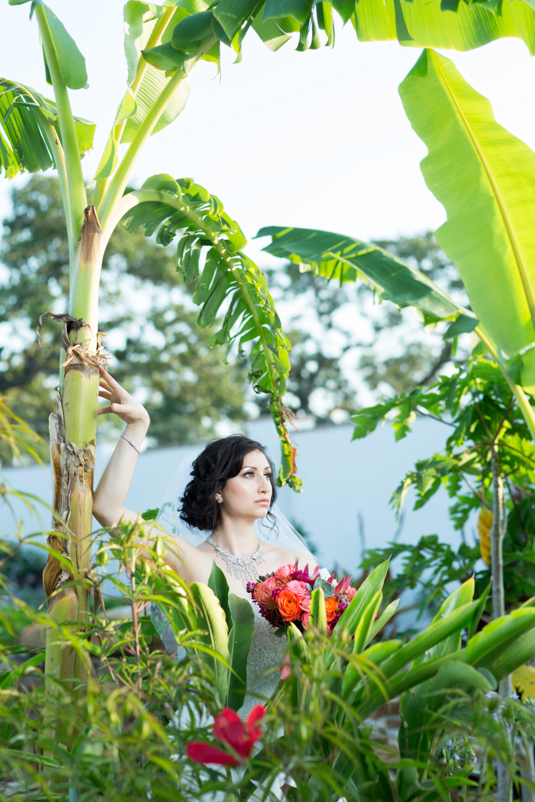 Bride in a white dress and veil holding a colorful bouquet among tropical plants and banana leaves.