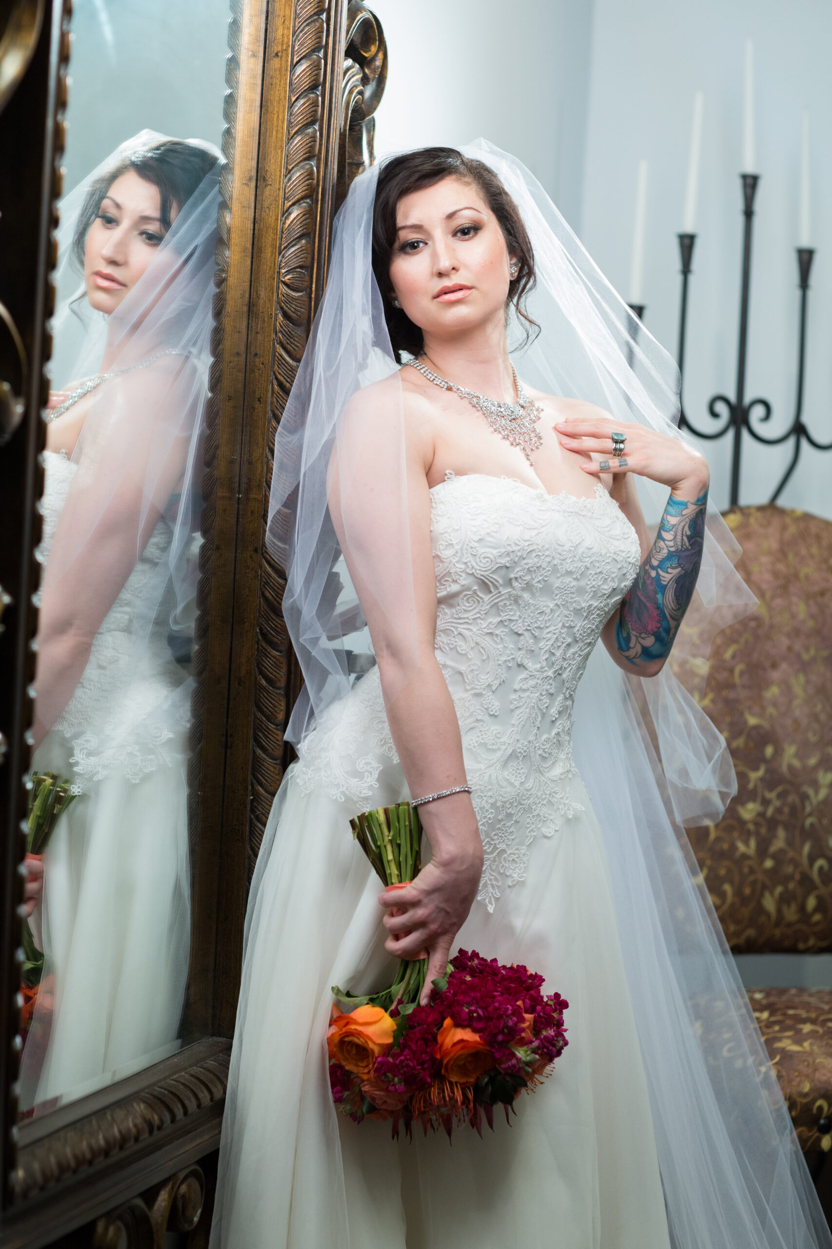 Bride in a lace wedding dress and veil holding an orange and burgundy bouquet beside an ornate mirror.