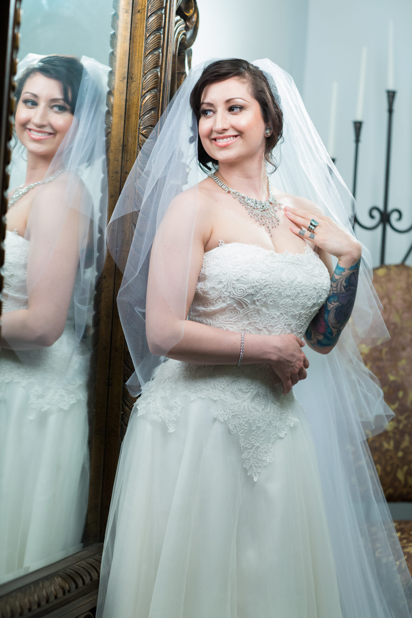 Jessica in a lace wedding dress and veil standing beside an ornate mirror, with her reflection visible.