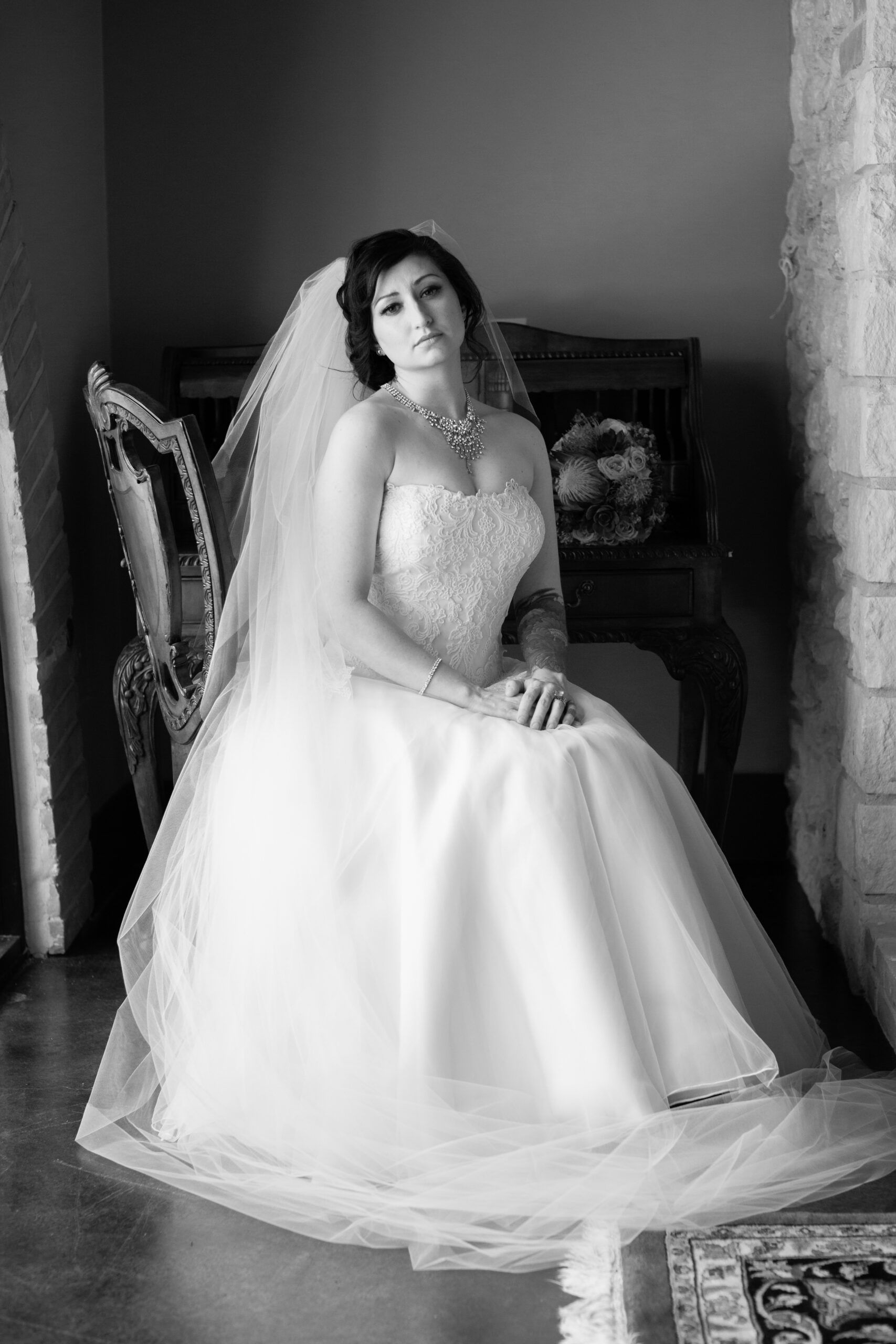 Black-and-white portrait of Jessica seated in a lace wedding gown and long veil beside a stone fireplace.