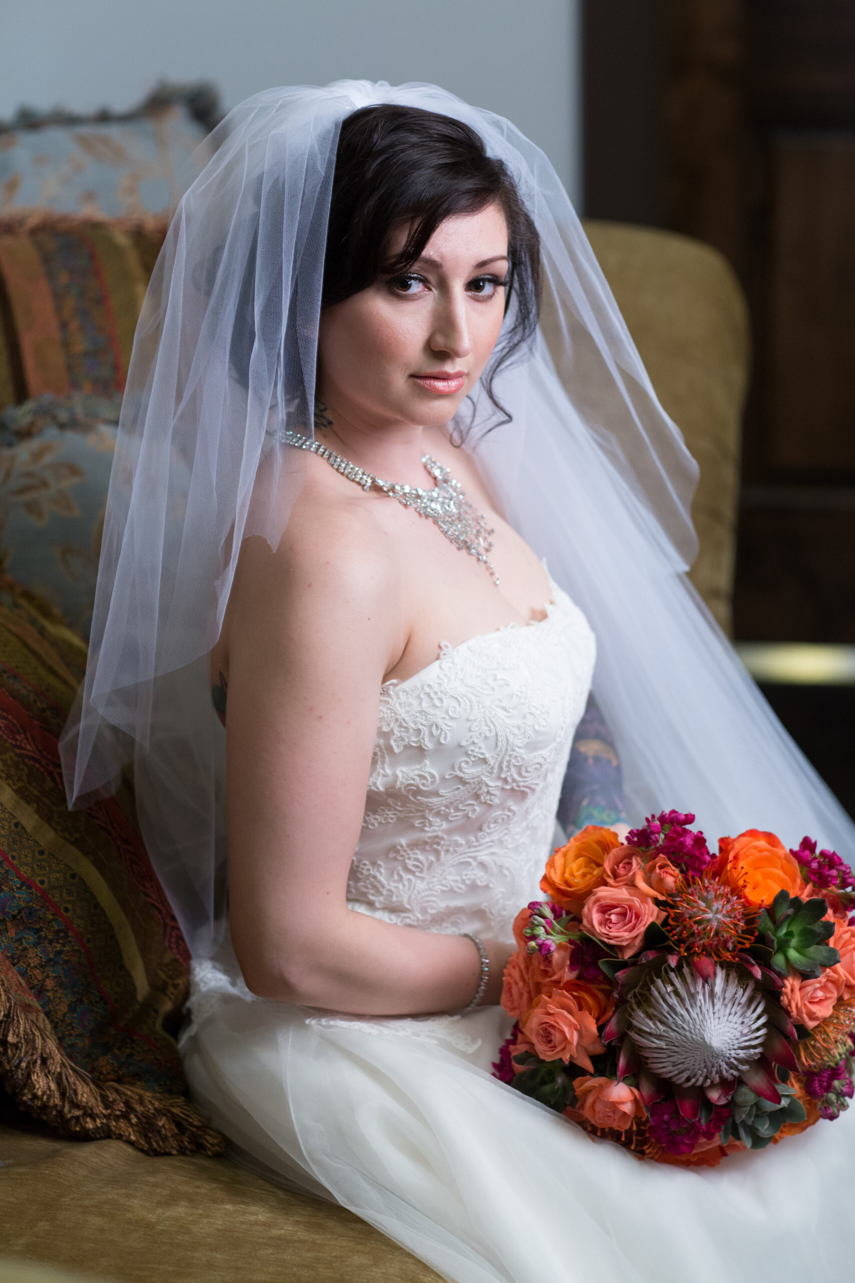 Bride Jessica seated indoors in a lace wedding dress and veil, holding a colorful bouquet.