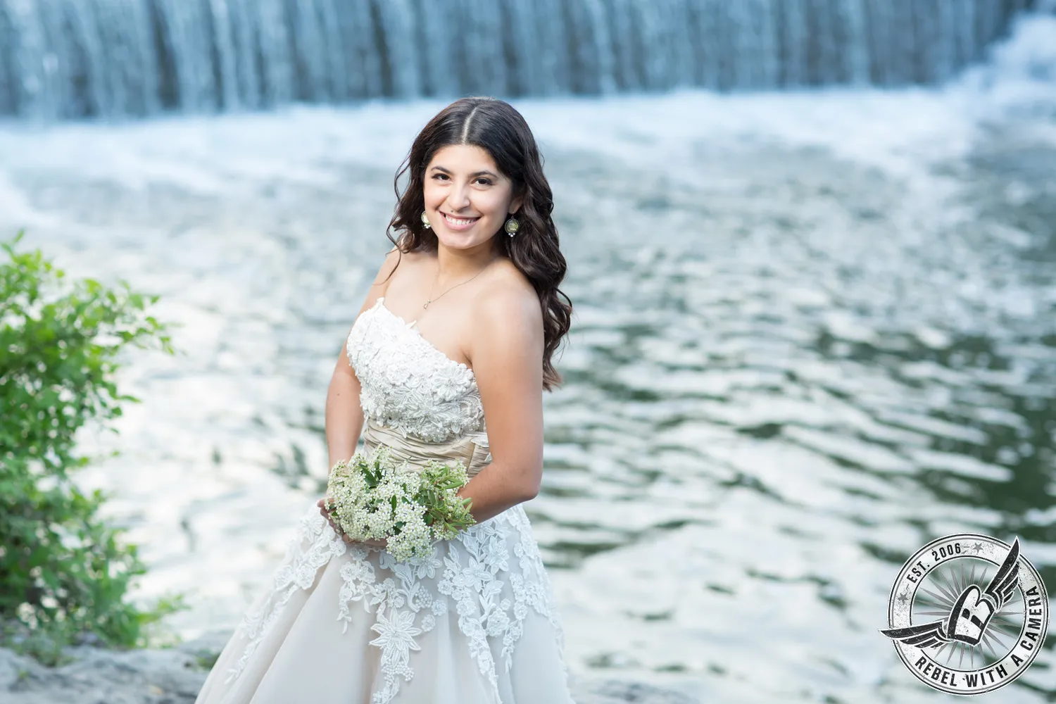 Bride in a lace wedding dress holding a bouquet beside a lake