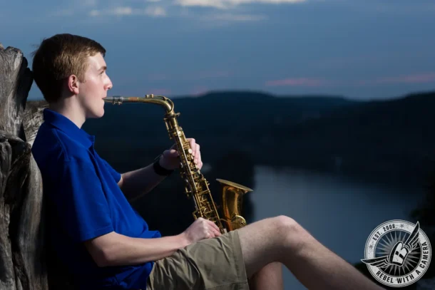 Johnathan sits beside a weathered wooden railing and plays a saxophone, with a river and dark hills in the background.