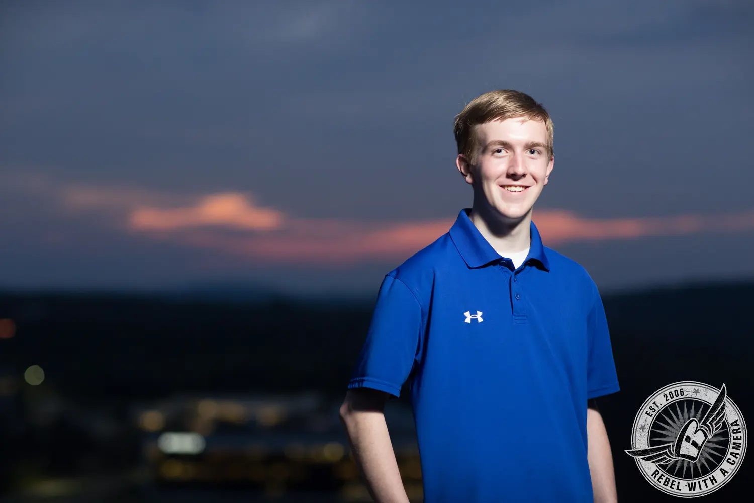 Johnathan wearing a blue polo shirt in an evening portrait with a dark cloudy sky and blurred city lights behind him.