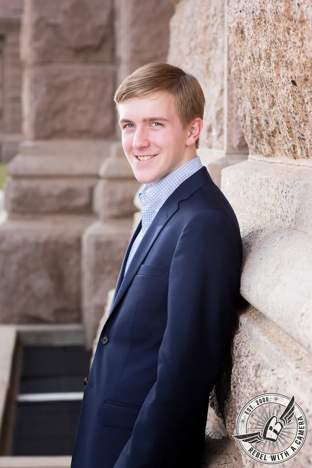 Johnathan in a navy suit jacket and checkered shirt leaning against a stone wall at the Texas State Capitol.