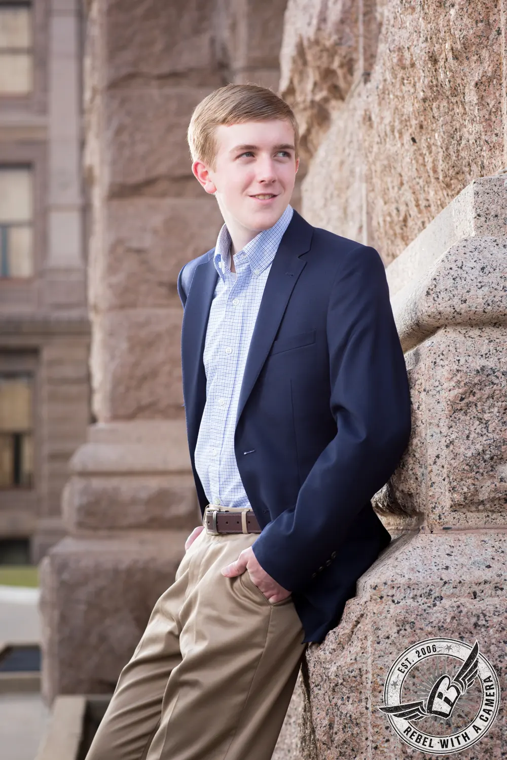 Johnathan in a navy blazer and khaki pants leaning against a stone wall at the Texas State Capitol.