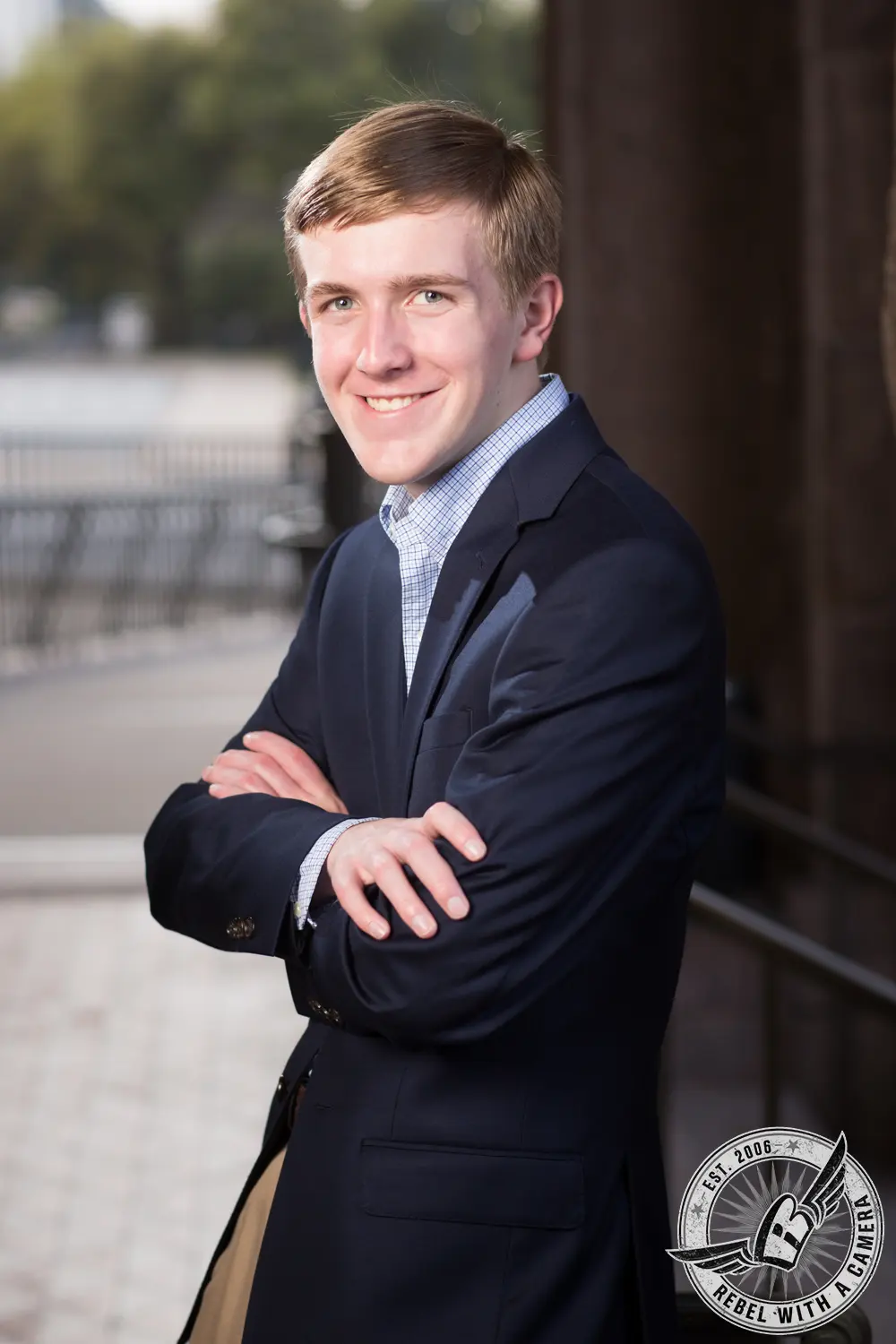 Johnathan stands with his arms crossed in a navy blazer and checkered shirt beside a dark stone wall.