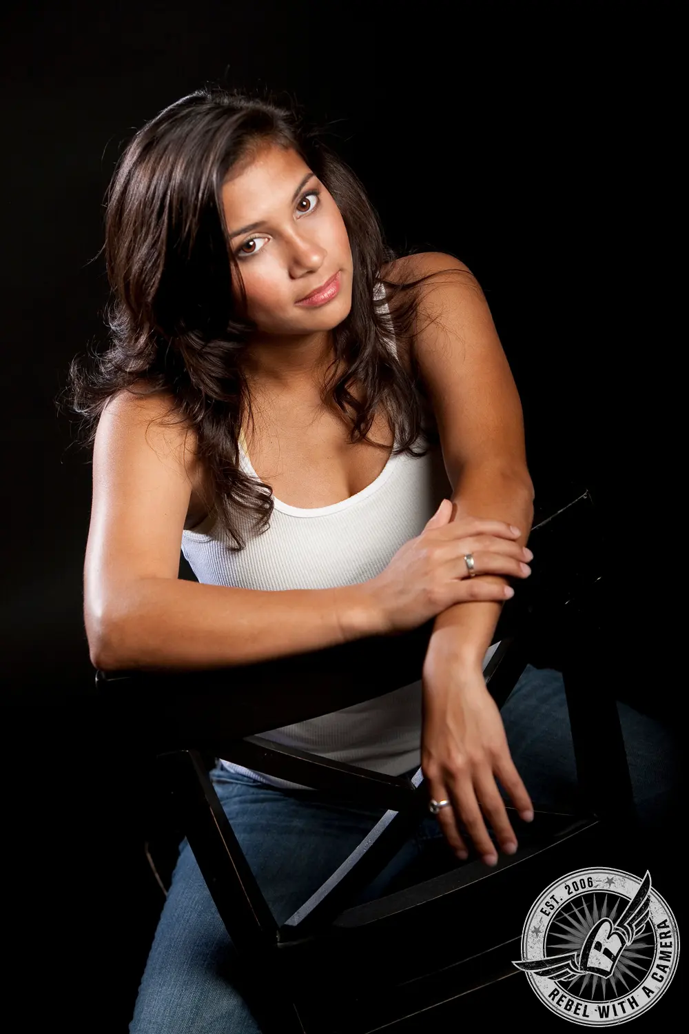 Katlyn seated backward on a black chair against a black studio background, wearing a white tank top and jeans.