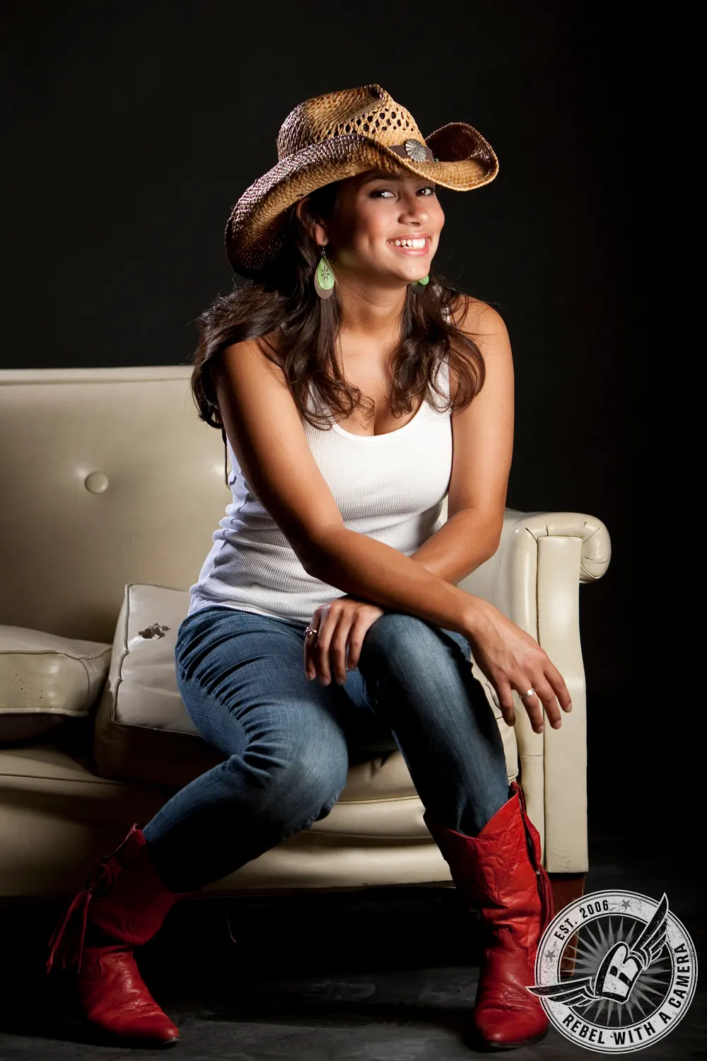 Katlyn sits on a cream leather chair in a studio, wearing a straw cowboy hat, white tank top, jeans, and red boots.