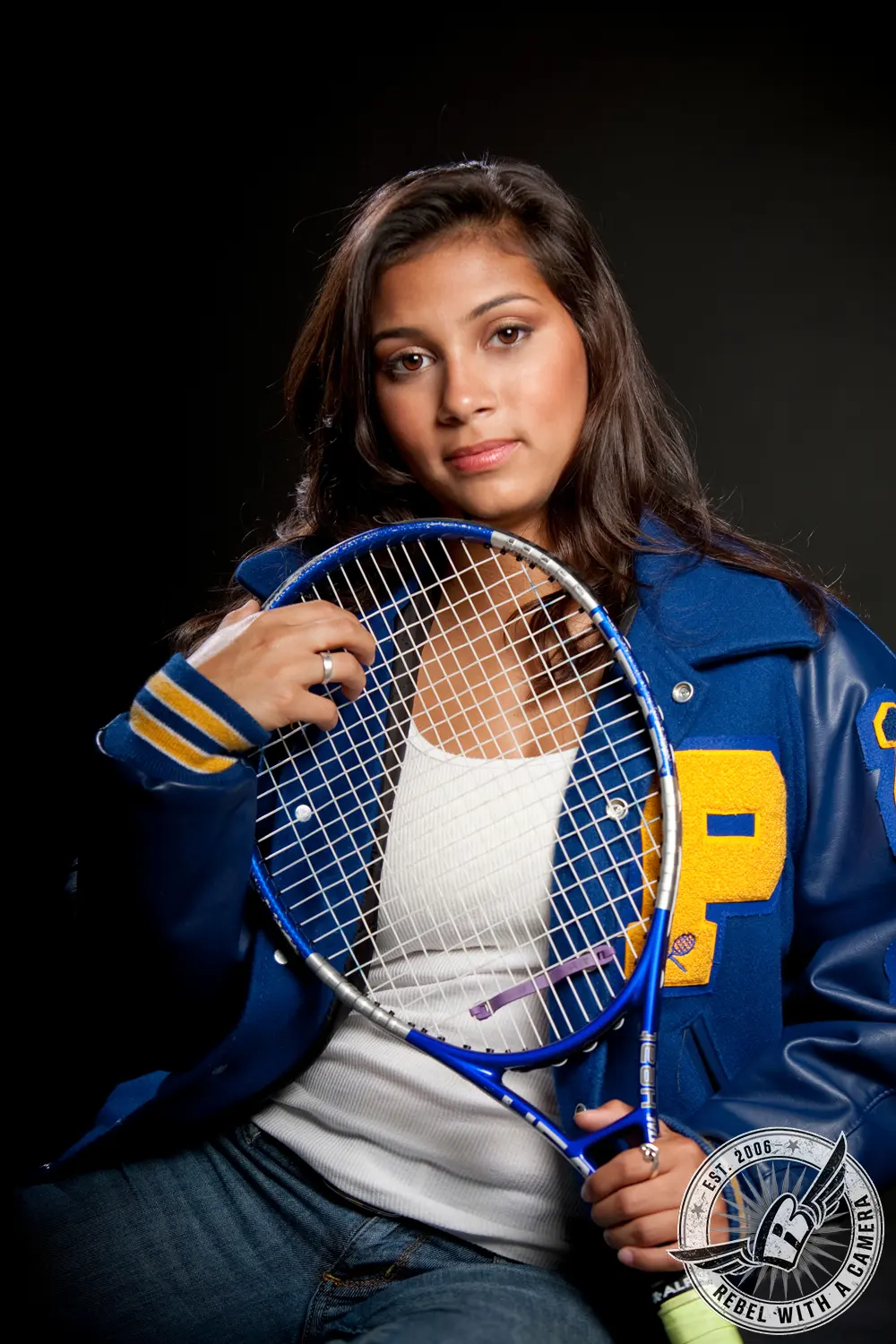 Katlyn holds a blue tennis racket while wearing a blue and gold letterman jacket against a dark studio background.