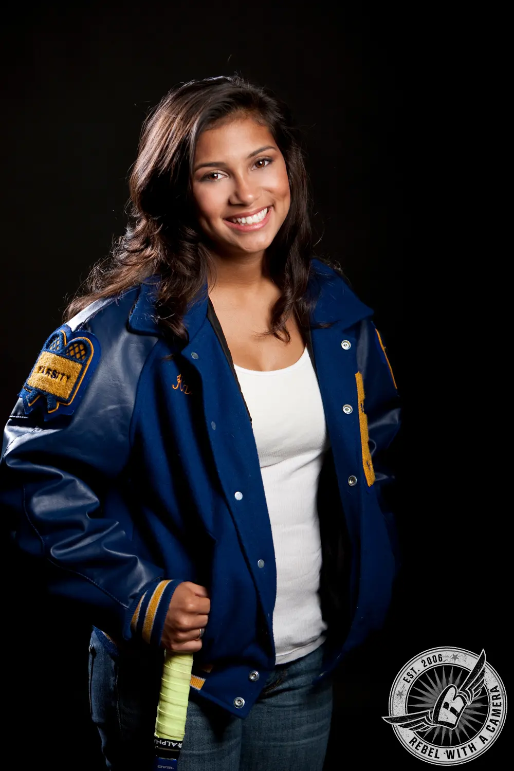 Studio portrait of Katlyn smiling in a blue tennis letter jacket while holding a tennis racket against a black background.