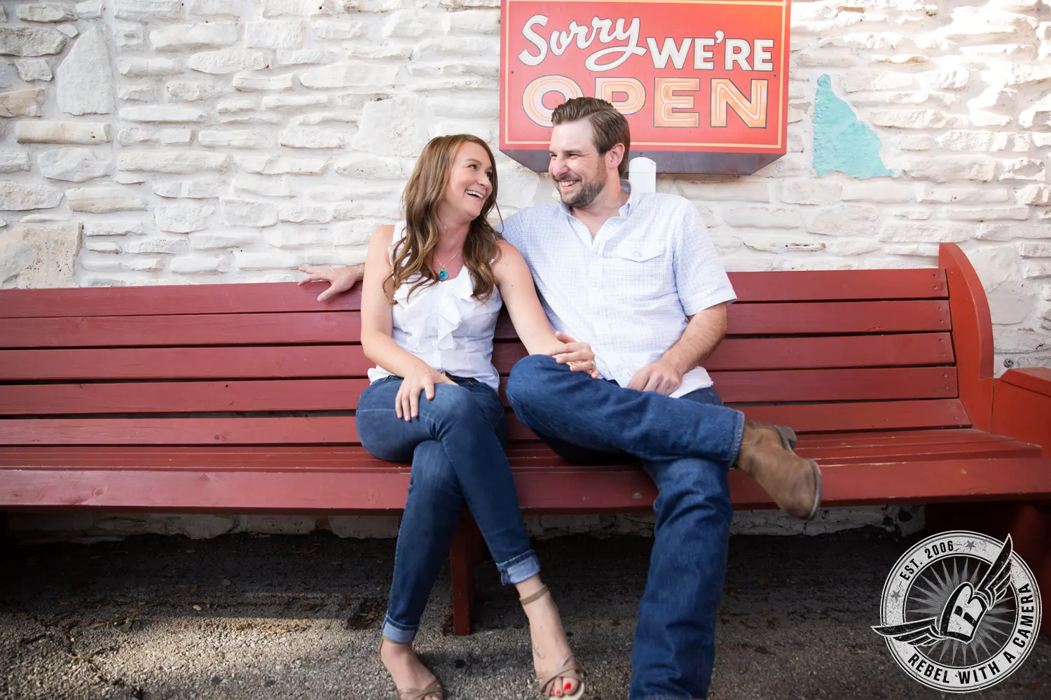 Couple laughing together on a red bench beneath an open sign in Austin, TX.