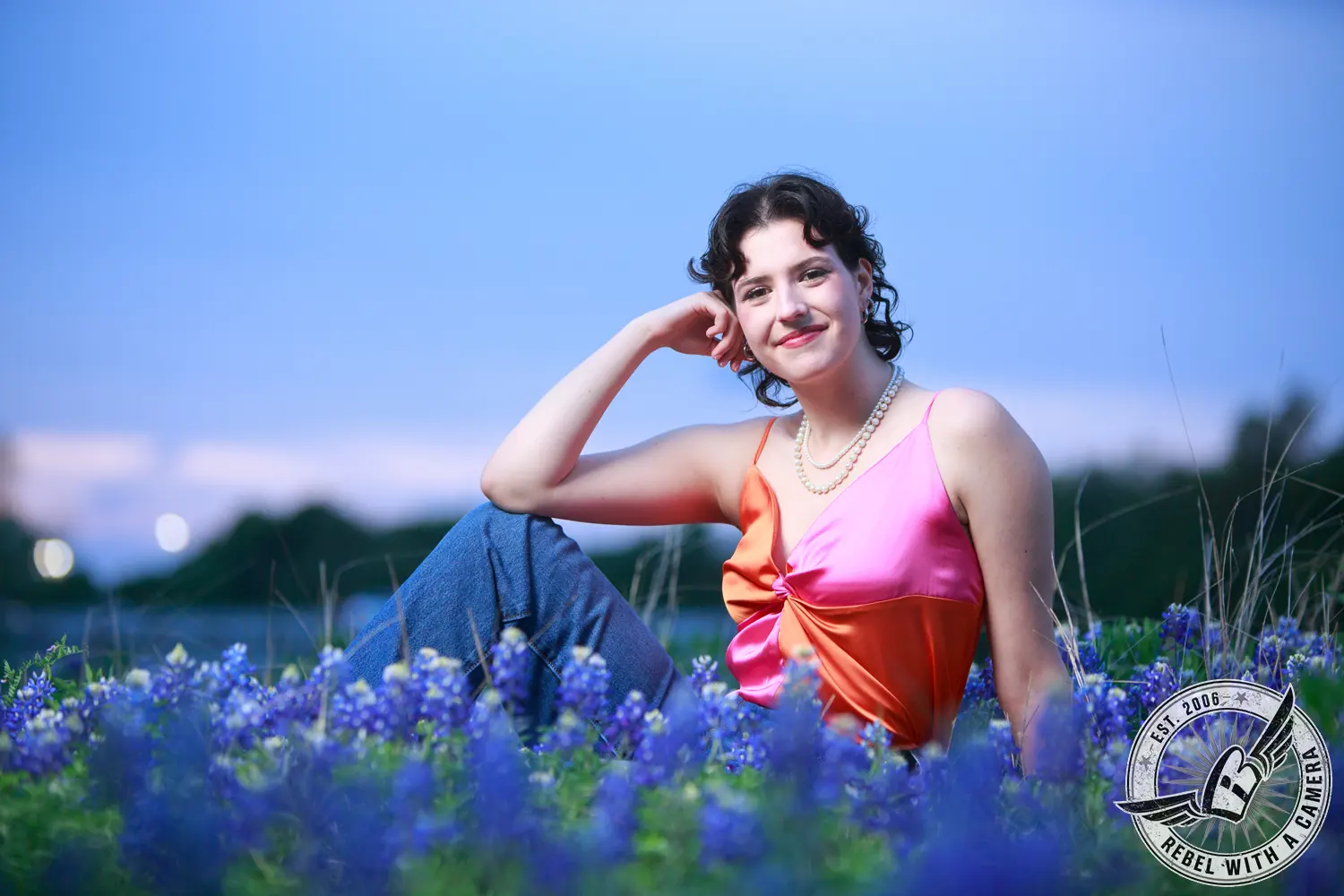 Liz seated among bluebonnets in a pink and orange top, jeans, and layered pearl necklaces.