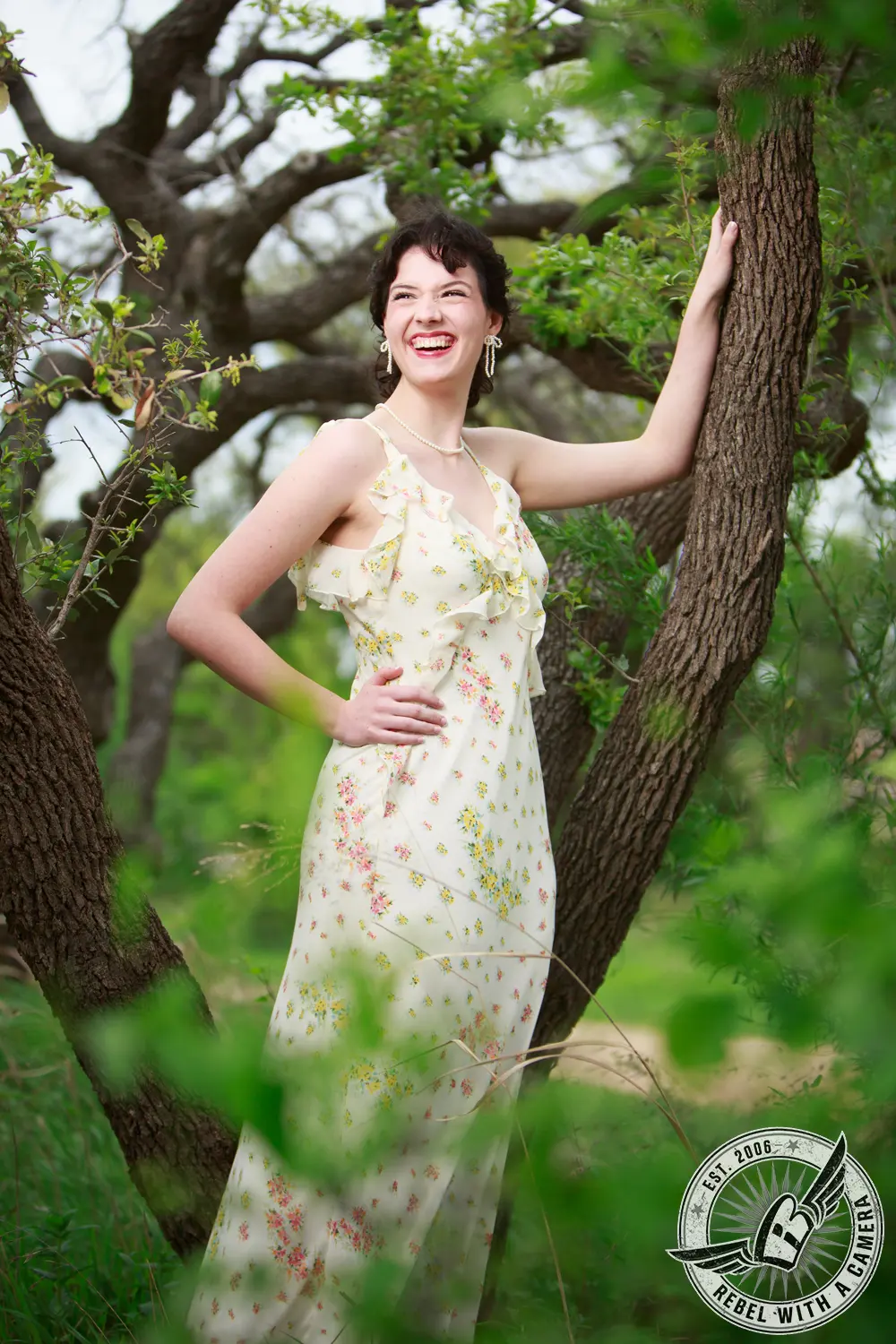 Liz smiling beside a tree in a floral dress, surrounded by green foliage.