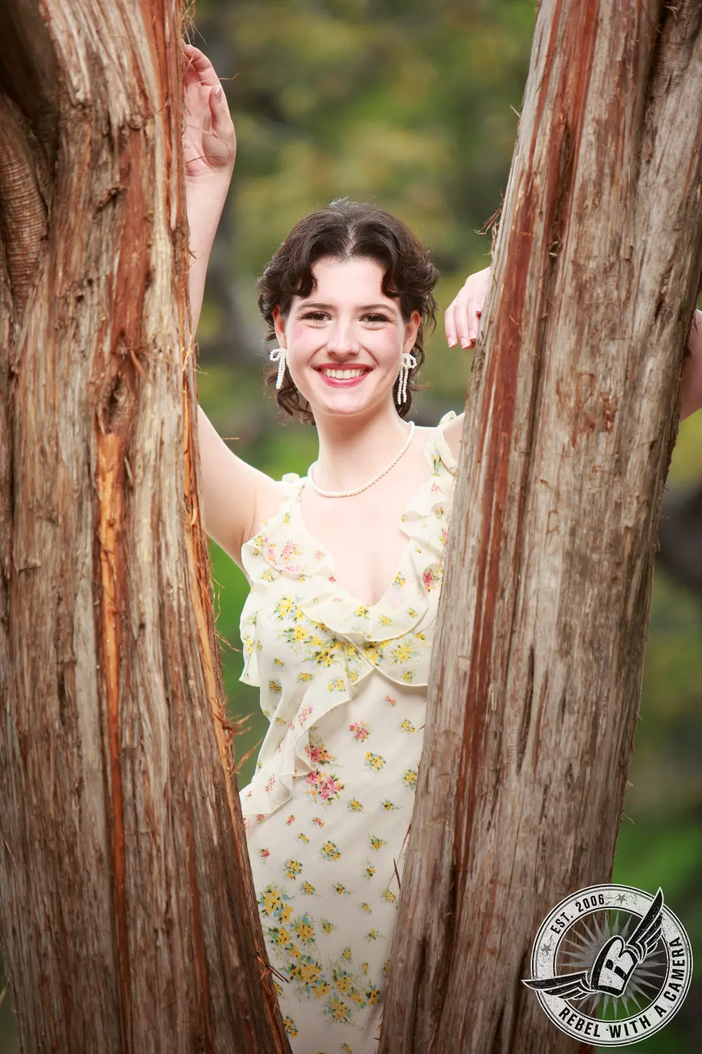 Liz smiles between two textured tree trunks while wearing a cream floral dress.