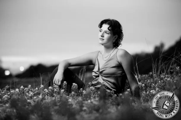 Black-and-white portrait of Liz seated among bluebonnets and tall grass, wearing a sleeveless dress and layered pearl necklace.