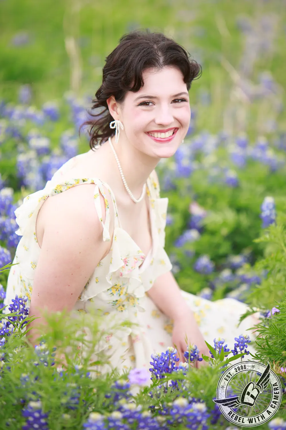 Liz sitting among bluebonnets in a light floral dress and pearl jewelry.