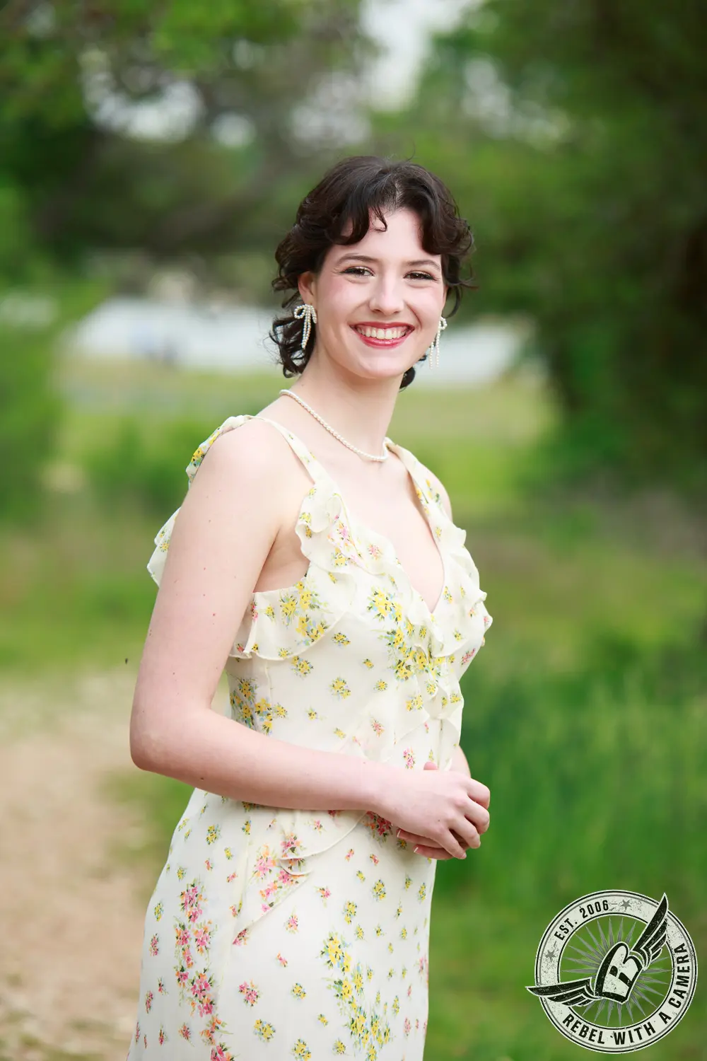 Liz standing outdoors in a light floral dress with pearl jewelry and bow-shaped earrings.