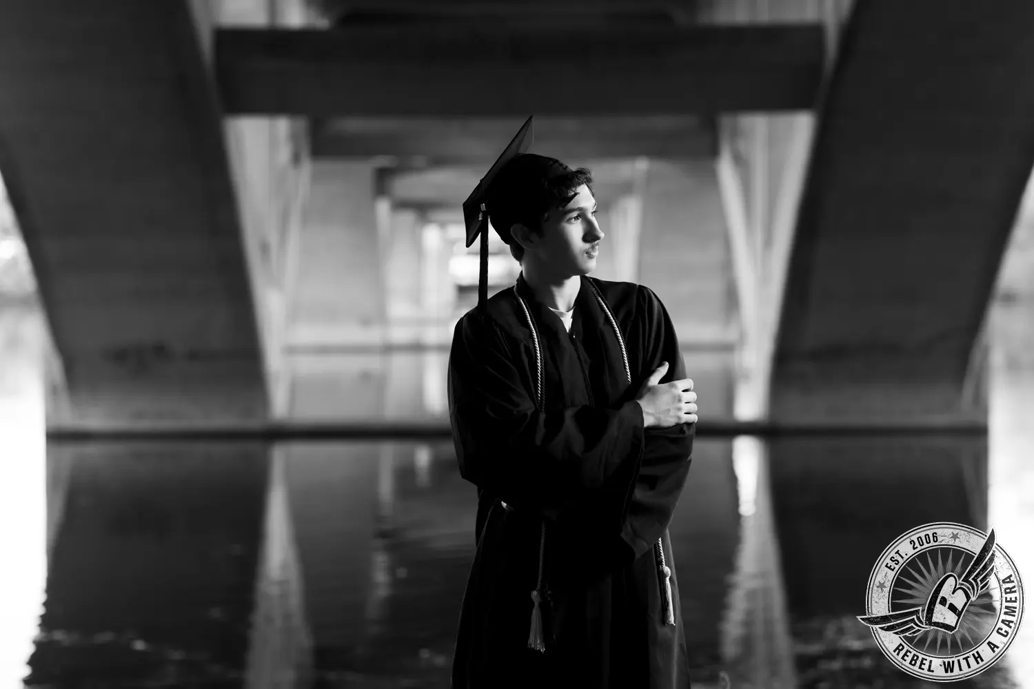 Black-and-white portrait of Nathan in a graduation cap and gown standing beneath a concrete bridge beside the water.
