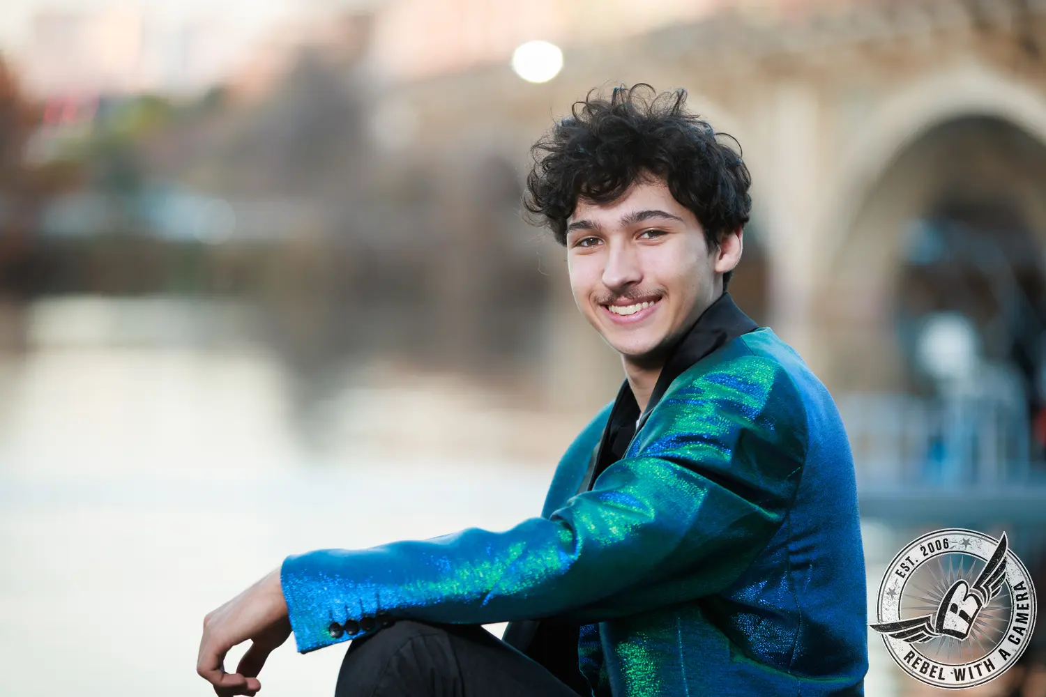 Nathan seated outdoors in a shimmering blue-green jacket, smiling toward the camera.