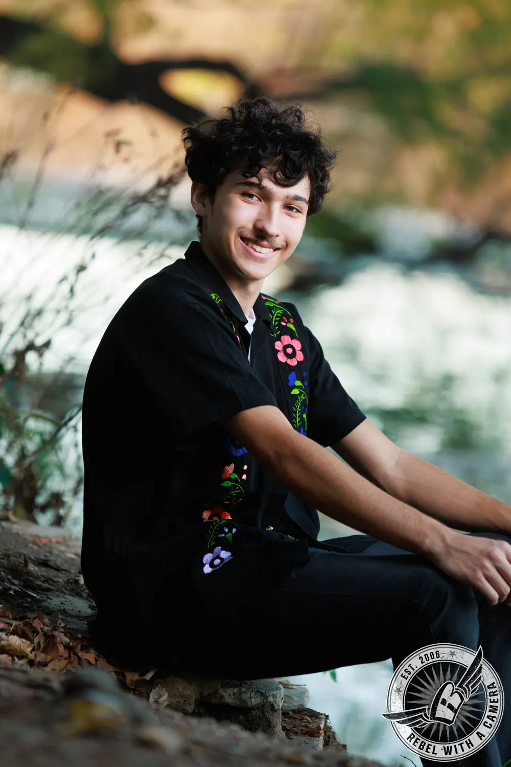Nathan seated on a stone ledge beside the water, wearing a black shirt with colorful floral embroidery.